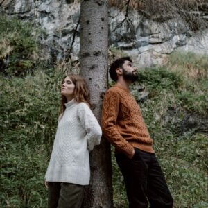 Home A couple stands back to back against a tree in an autumn forest setting.