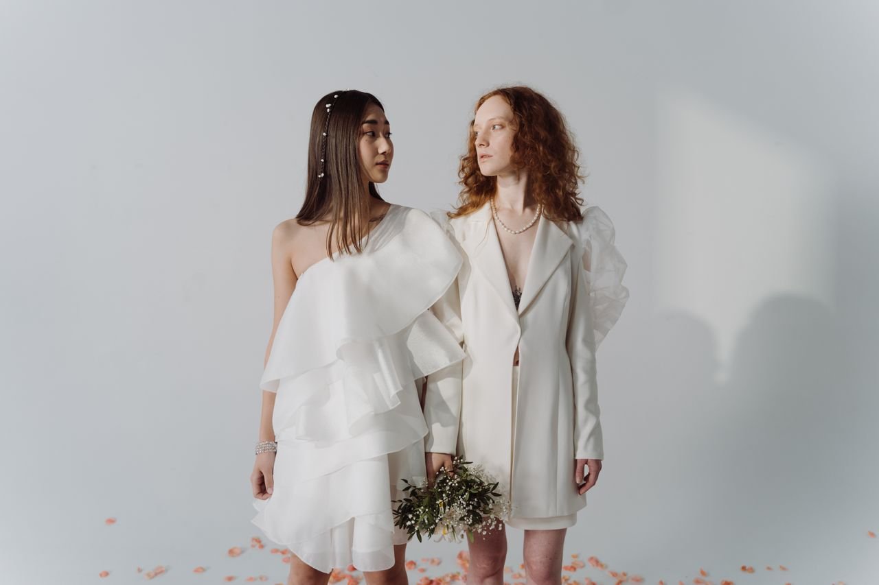 Home A stylish same-sex couple in white attire holding a bouquet during a studio wedding photoshoot.